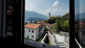 Une vue depuis une fenêtre donnant sur une ville, avec un clocher d’église imposant, des bâtiments en pierre et un paysage montagneux en arrière-plan.