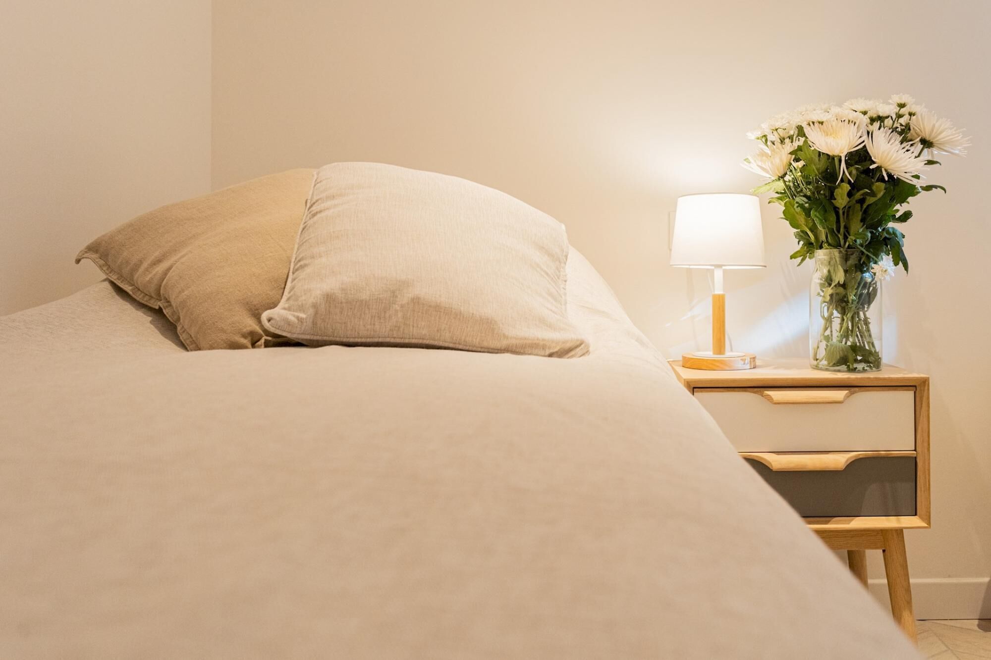 A bed with a beige blanket and pillows, a bedside table with a lamp and a vase of flowers.