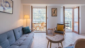 A modern living room with a grey sofa, a small round table, and a chair with a yellow cushion. There is a large window with a view of a lake and trees. - Co-e405-regi74a1 - Your Private Harbour Nearby George Clooney (Carate Urio)