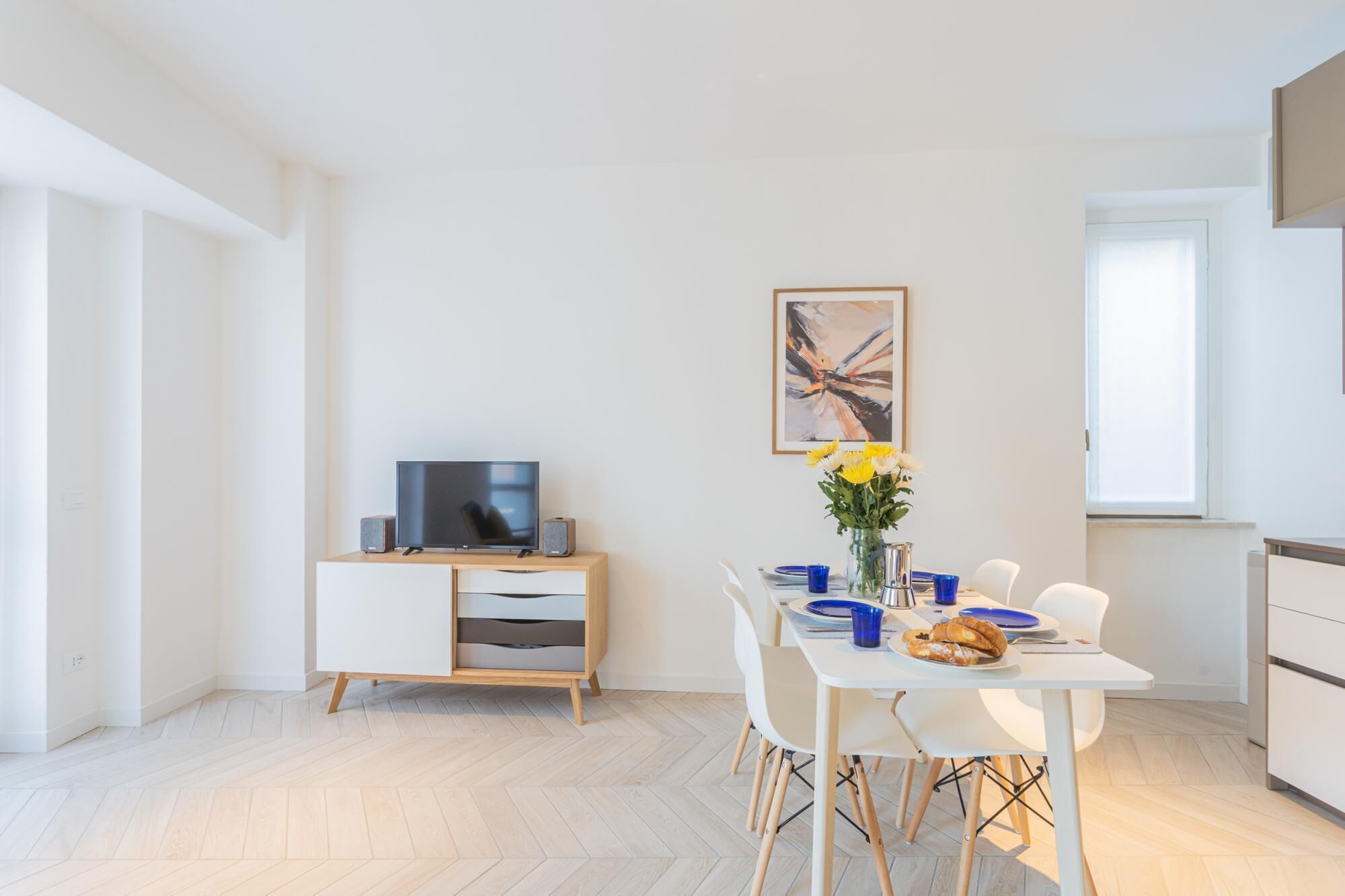Une cuisine moderne avec un coin repas, un téléviseur à écran plat posé sur un meuble en bois, et une table dressée pour deux personnes, agrémentée d’un vase de fleurs.