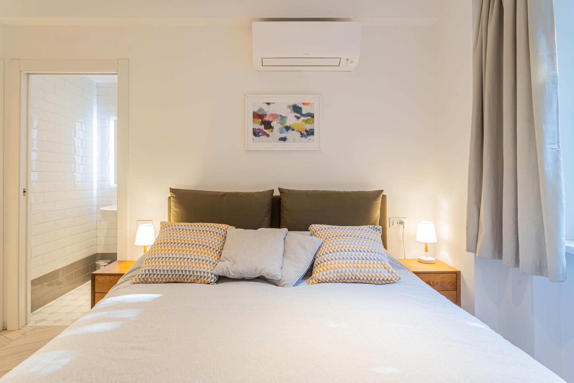 A hotel room with a large bed, two bedside tables with lamps, a wall-mounted air conditioner, and a window with curtains.