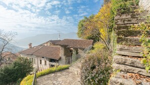 A stone staircase leading to a traditional house with a tiled roof, surrounded by greenery and overlooking a mountainous landscape. - Co-d462-mpal3at - Villa Sunset Tower (Faggeto Lario)