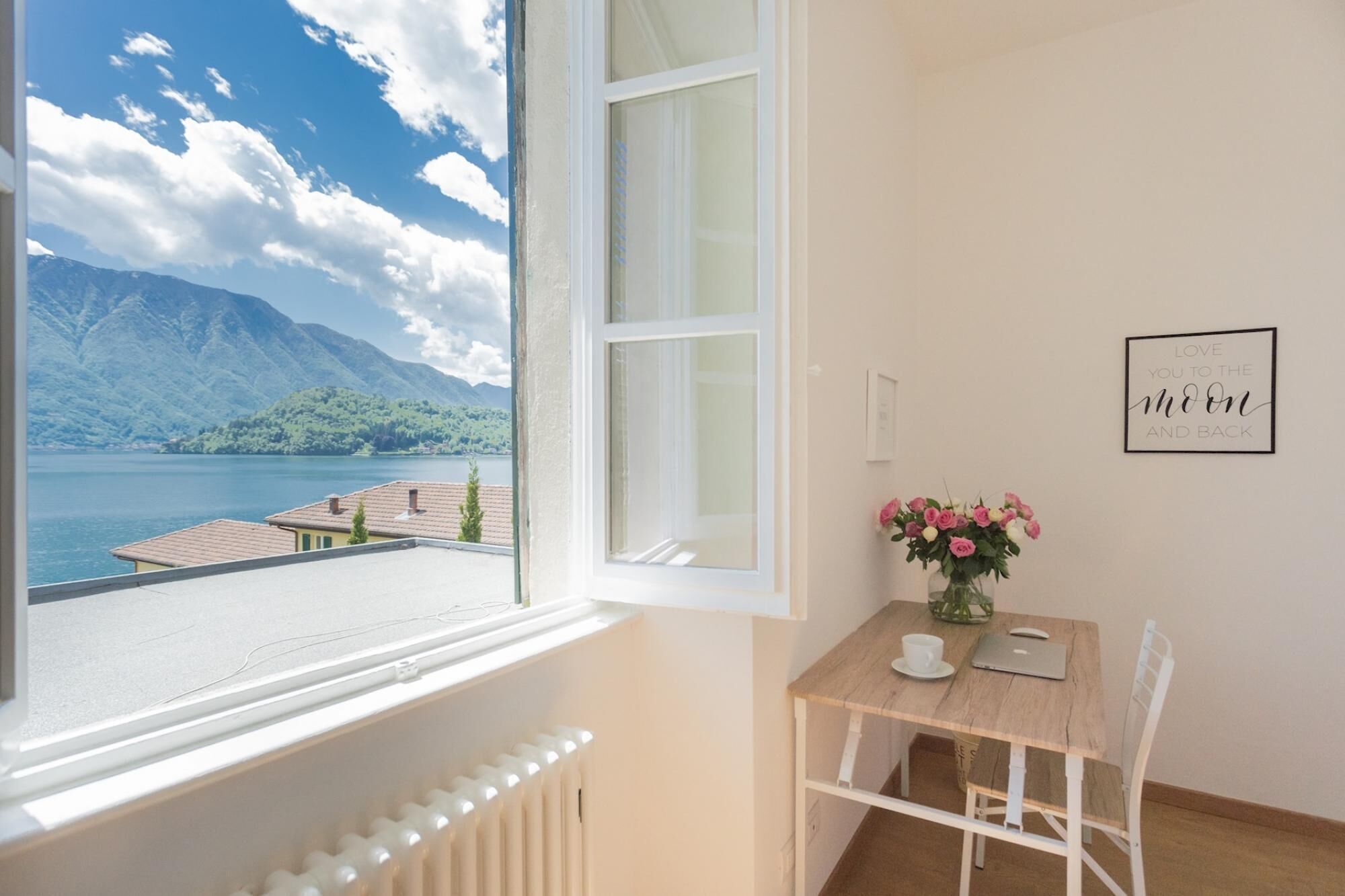 A room with a view of a lake and mountains, a wooden table with a laptop, a cup, and a vase of flowers.