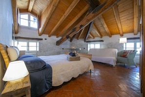 Une chambre à coucher au style rustique, avec un plafond en bois apparent, un grand lit recouvert d’une courtepointe blanche, une table de chevet avec une lampe et une chaise agrémentée d’un coussin.
