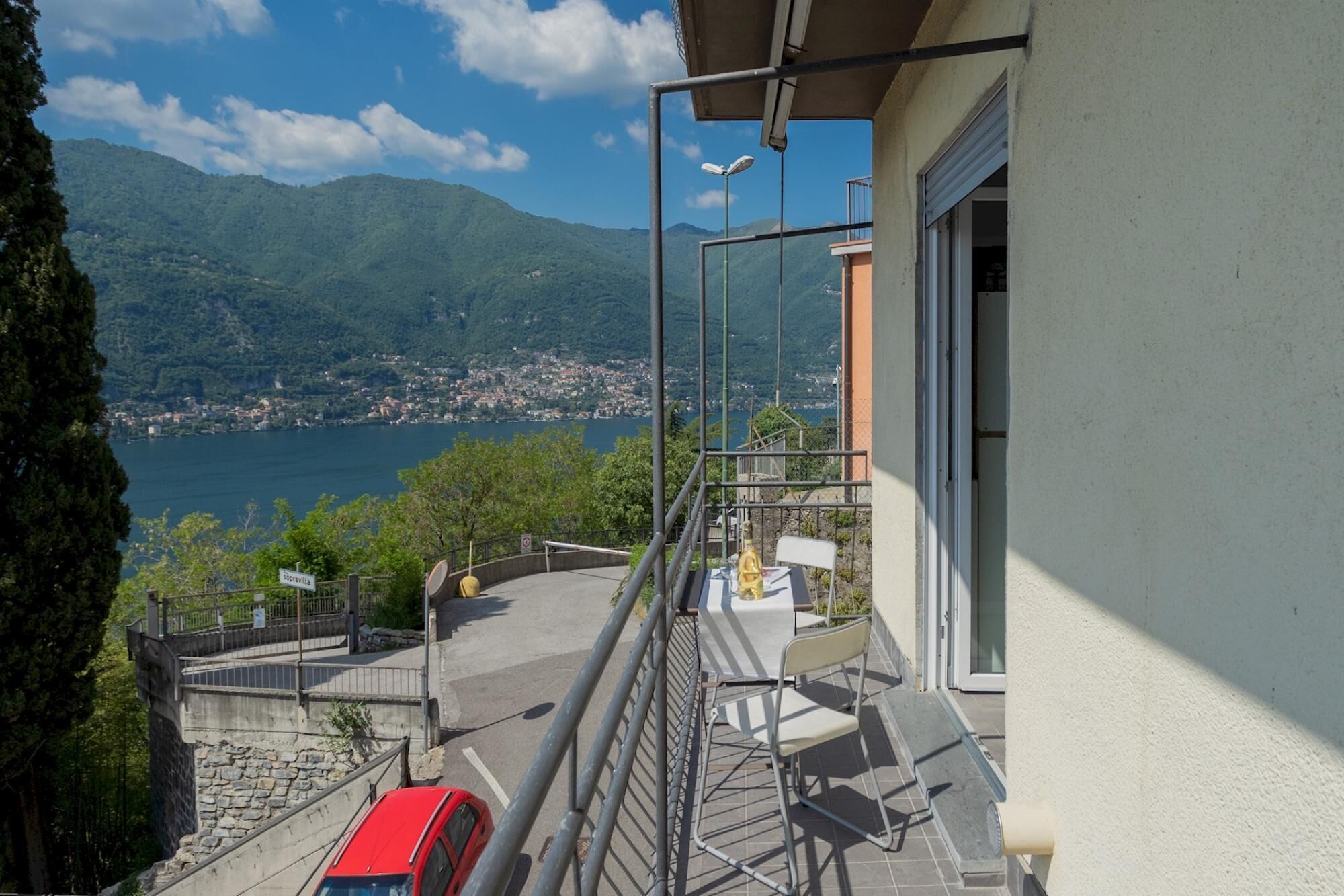 A balcony with a view of a lake and mountains, a red car parked below, and a chair on the balcony.
