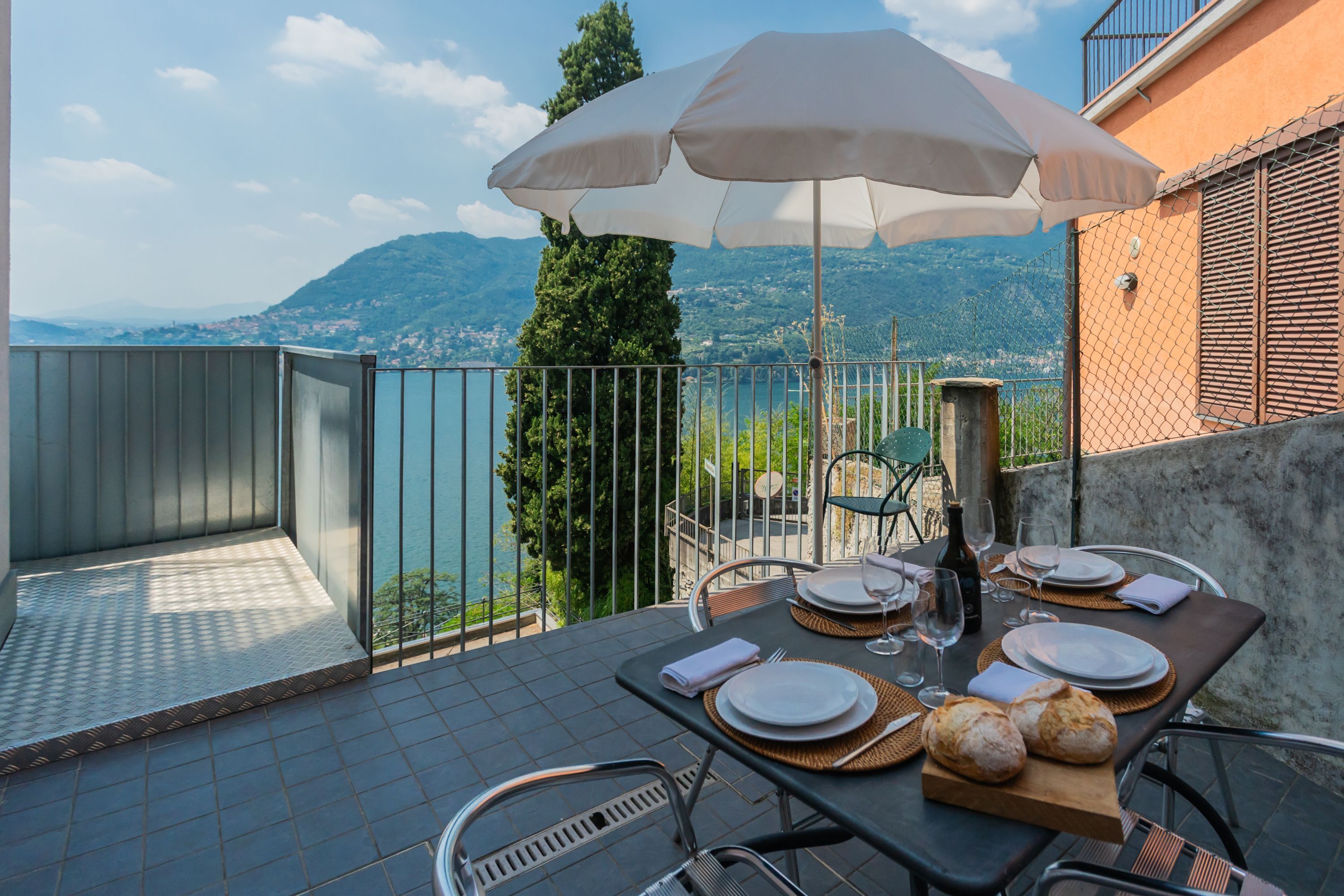 A dining table set with plates, glasses, and bread on a balcony with a view of mountains and a body of water.