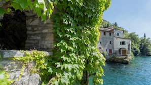 A stone building with a red door, partially covered by green ivy, adjacent to a body of water.