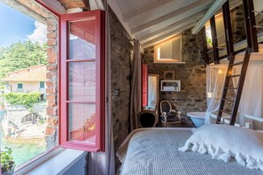 A bedroom with a bed, a window with a red shutter, a stone wall, a wooden ladder, and a view of a house and greenery outside. - La Civera - Nesso Lake Como - Co-f877-pont41ar (Nesso)