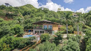 Ein modernes, zweistöckiges Haus mit einem großen Balkon, umgeben von üppiger Vegetation und einem Schwimmbecken.