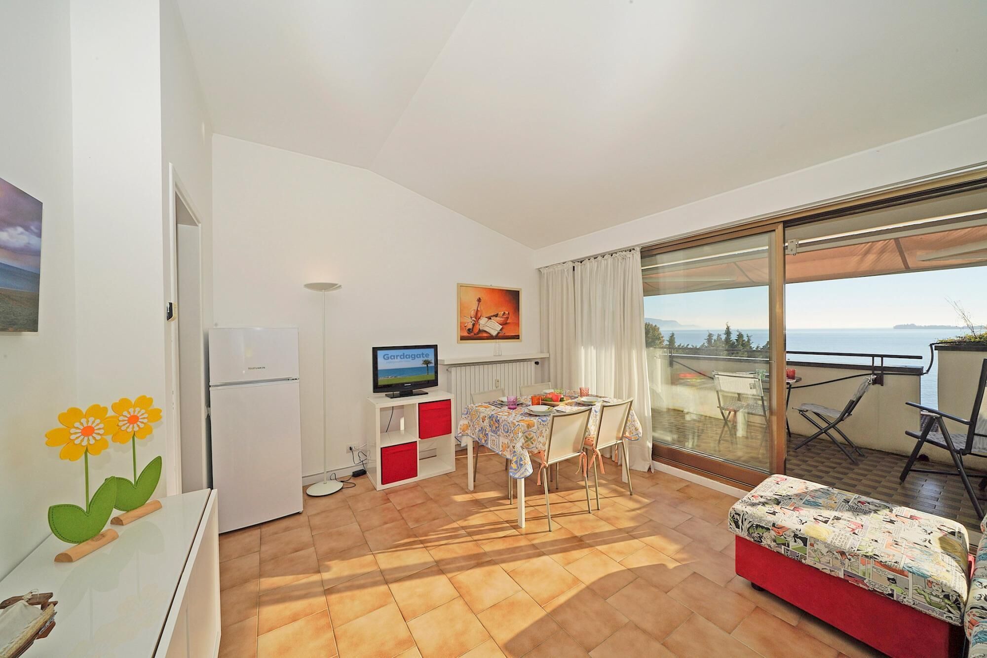 A compact living space with a dining area, a television, a refrigerator, and a balcony with a view of the sea.
