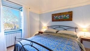 Ein Schlafzimmer mit einem Bett, zwei Nachttischlampen, einem Fenster mit Vorhängen und einem an der Wand hängenden Kunstwerk.