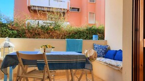 En balkon med et bord, stole og en sofa. Der er en potteplante på bordet og en dekorativ lygte. Balkonen har udsigt til en bygning med balkoner og en hæk.