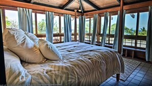 Una cama grande con almohadas, vigas de madera y vista al océano a través de ventanas.