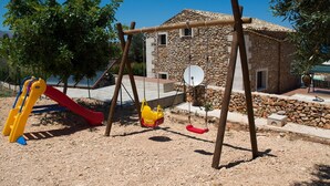 Ein Spielplatz mit Rutsche und Schaukeln, umgeben von einem Steinhaus und Bäumen.