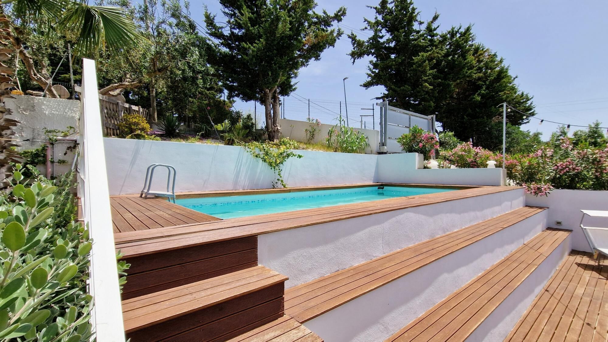 Una piscina con escalones de madera que la bordean, rodeada de vegetación y un muro blanco.