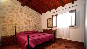 A bedroom with a stone-patterned wall, a wooden ceiling, a bed with a metal frame, a nightstand, a dresser, a mirror, and a window with curtains. - Dependance Desir e Piscina ad uso Esclusivo e Jac (Alcamo)