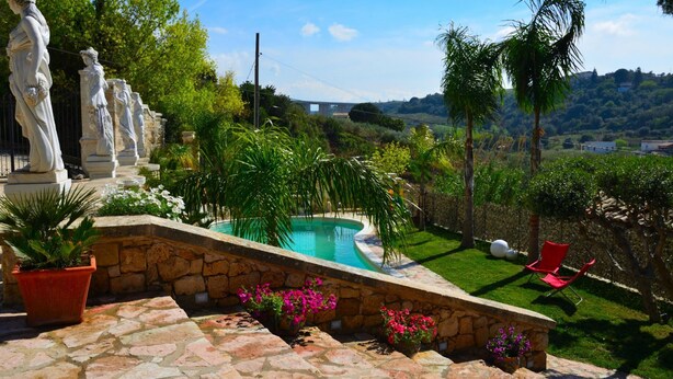 A landscaped garden with a swimming pool, stone statues, and a red chair. - Dependance Desir e Piscina ad uso Esclusivo e Jac (Alcamo)