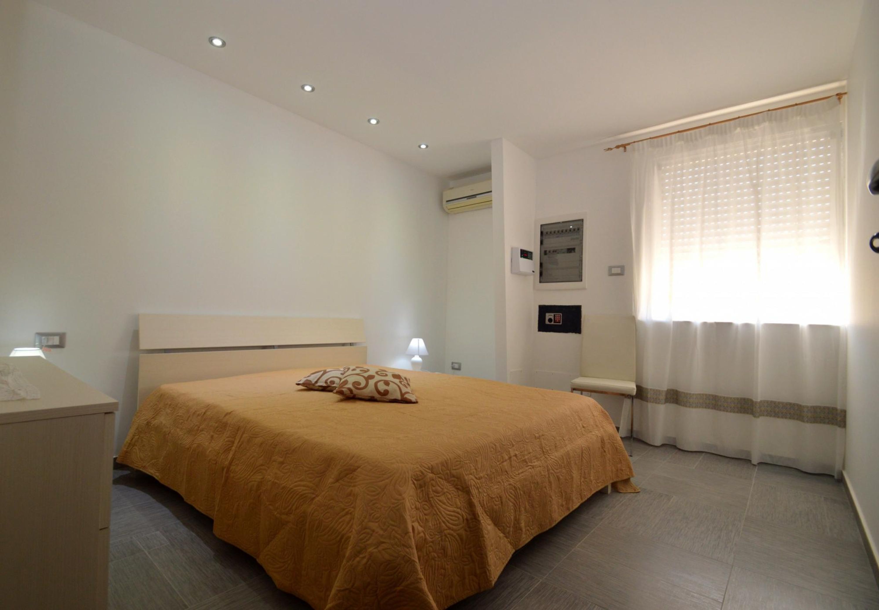 Ein Schlafzimmer mit Bett, Nachttisch, Lampe, Klimaanlage und einem mit Vorhängen versehenen Fenster.
