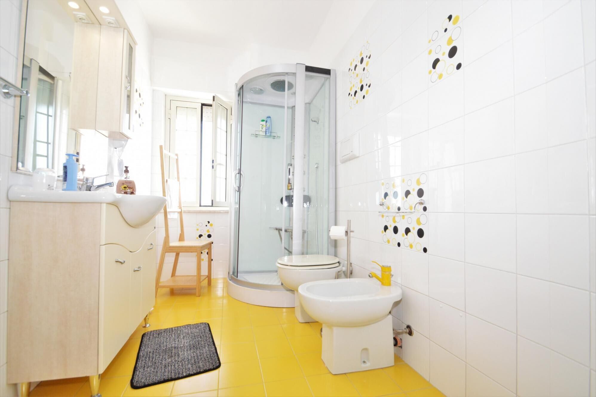 Un baño moderno con piso amarillo, azulejos blancos, cabina de ducha de vidrio, inodoro y lavamanos con espejo arriba.