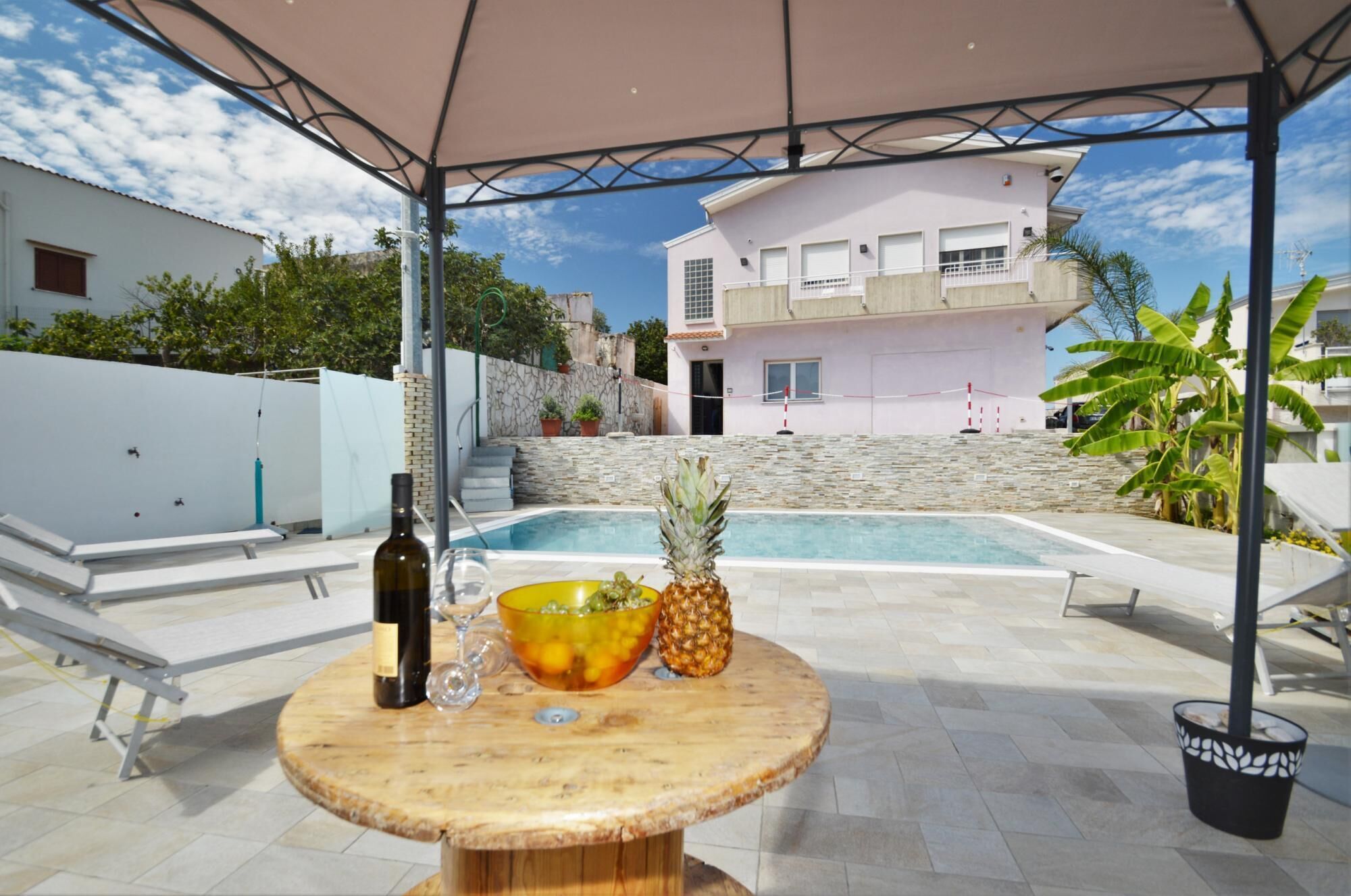 Un espace aménagé au bord de la piscine, avec une table sur laquelle se trouvent une bouteille, un verre à vin et un saladier rempli de fruits.
