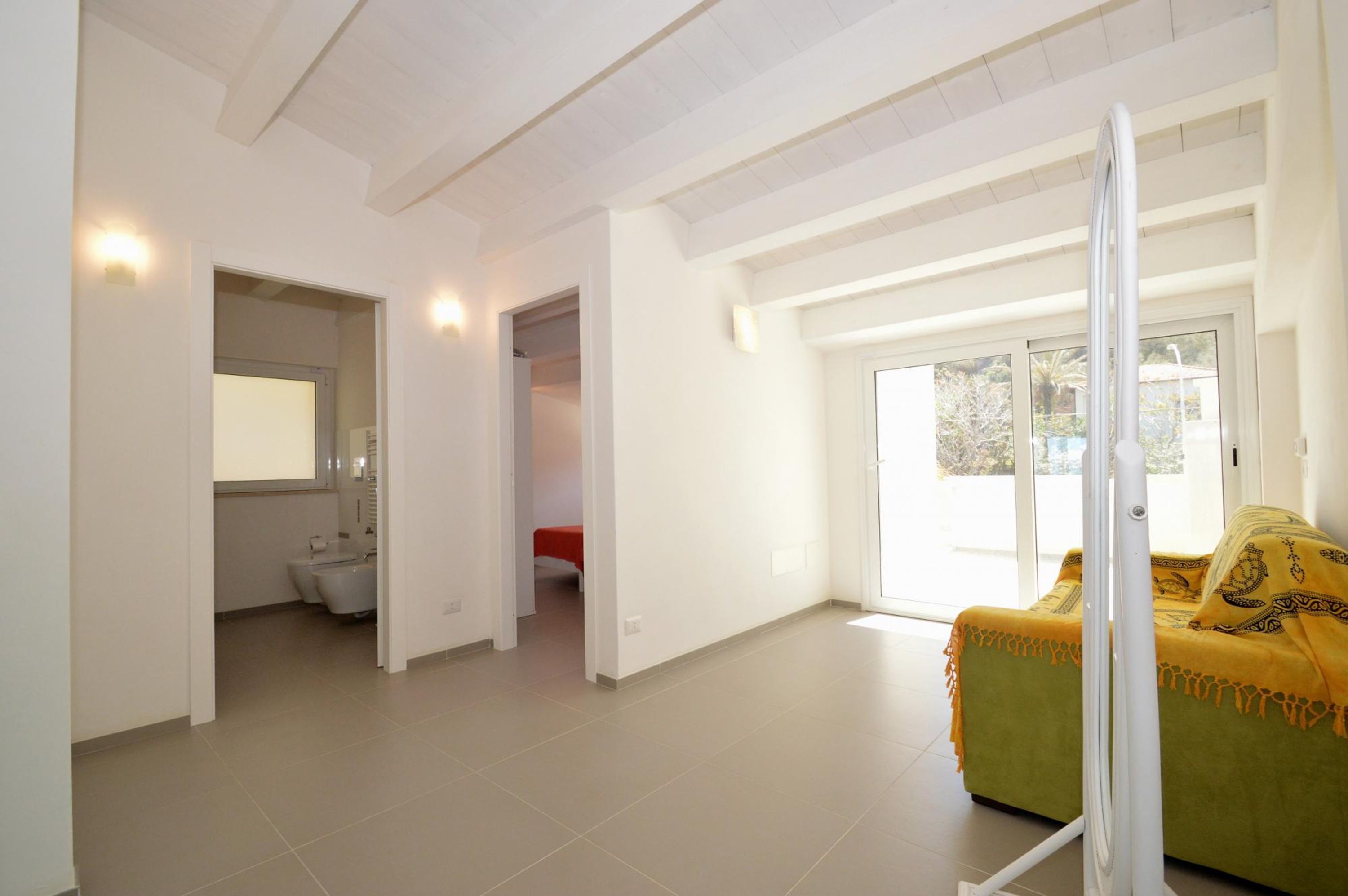 A spacious room with a large window, a bed with a yellow blanket, and a bathroom with a toilet and sink.