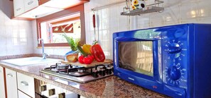 Une cuisine avec un micro-ondes bleu, une cuisinière à gaz et une fenêtre donnant sur un paysage verdoyant.