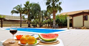 Une table au bord de la piscine avec un verre de vin rouge, deux oranges, une assiette de raisins et une tranche de pastèque.