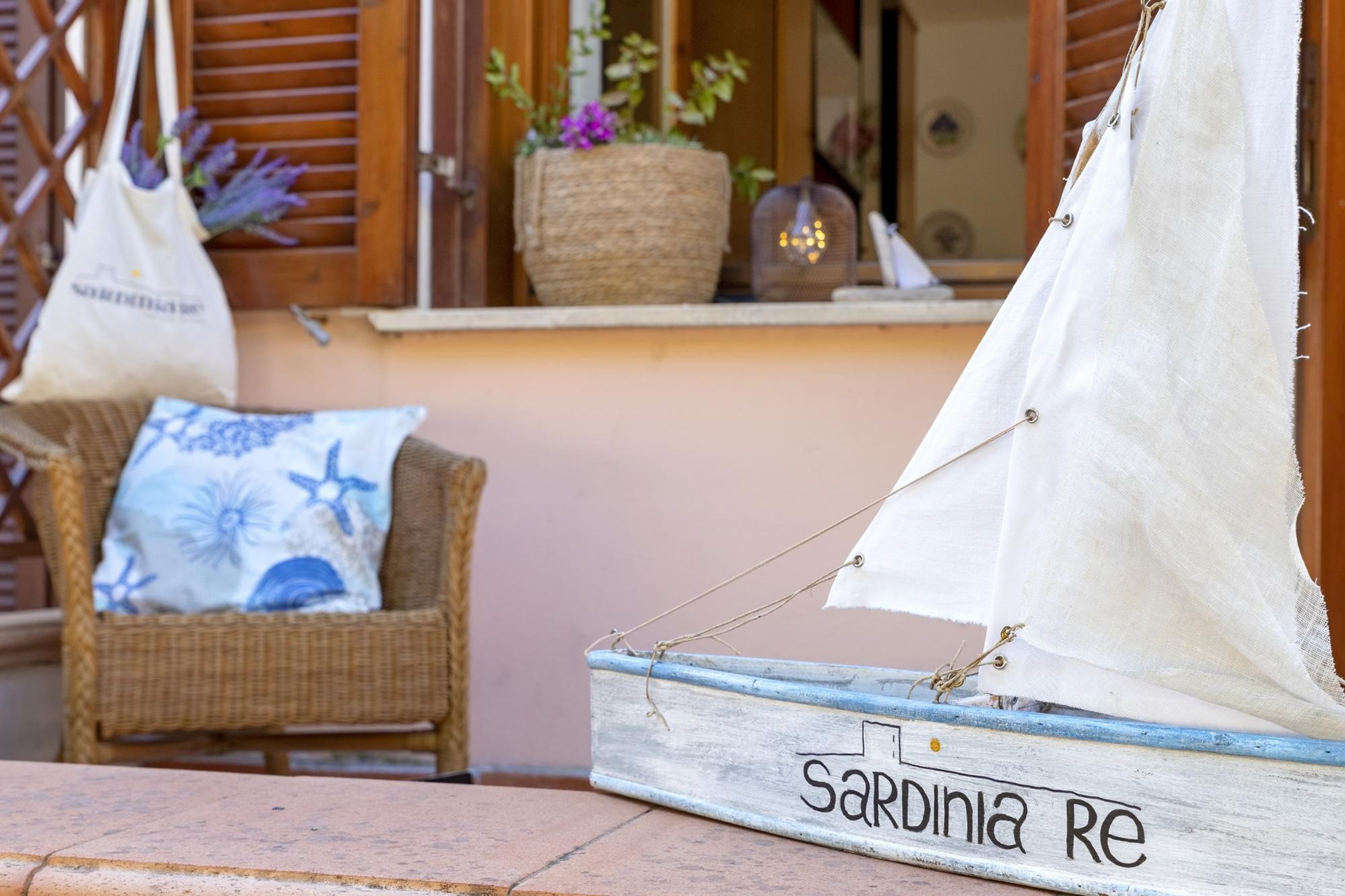 A rustic boat with 'Sardinia Re' written on it, a wicker chair with a starfish pillow, and a wooden window with shutters.