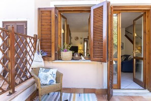 A wooden balcony with open shutters, a wicker chair with a cushion, and a small rug on the floor.