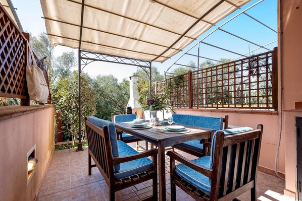 A covered outdoor patio with a table set for dining, surrounded by wooden chairs with blue cushions.