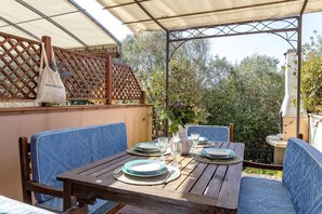 An outdoor dining area with a wooden table set for two, blue cushioned chairs, and a white vase with flowers.