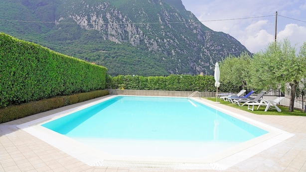 Una piscina rectangular con un bordeado de baldosas, rodeada por un seto bajo y una valla de metal. Al fondo, un paisaje montañoso con cielo despejado.