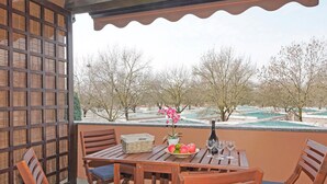 A wooden table with chairs, a basket, a plate of fruit, and wine glasses on a balcony with a view of trees and a pool. - Casa Rudi in Sirmione (Sirmione)