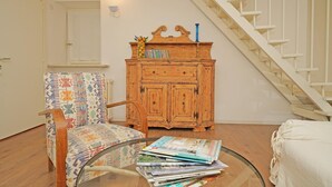 A living room with a wooden cabinet, a patterned armchair, a glass coffee table with magazines, and a staircase.
