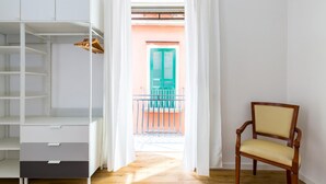 Une pièce avec un plancher en bois, une armoire blanche, une chaise et une vue sur un balcon aux volets verts.