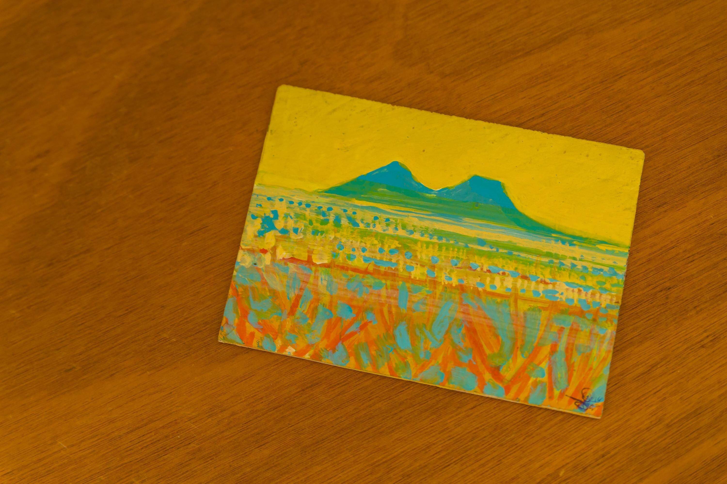 Une petite peinture carrée représentant un paysage de montagne et de champs, réalisée sur une surface en bois.