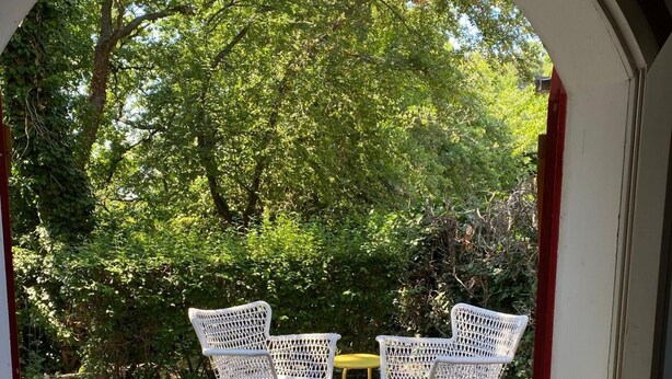Een terras buiten met twee witte rieten stoelen en een kleine gele tafel, omgeven door weelderige begroeiing en bomen.