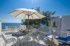A terrace with white metal chairs and tables, a large umbrella, and a view of the sea.