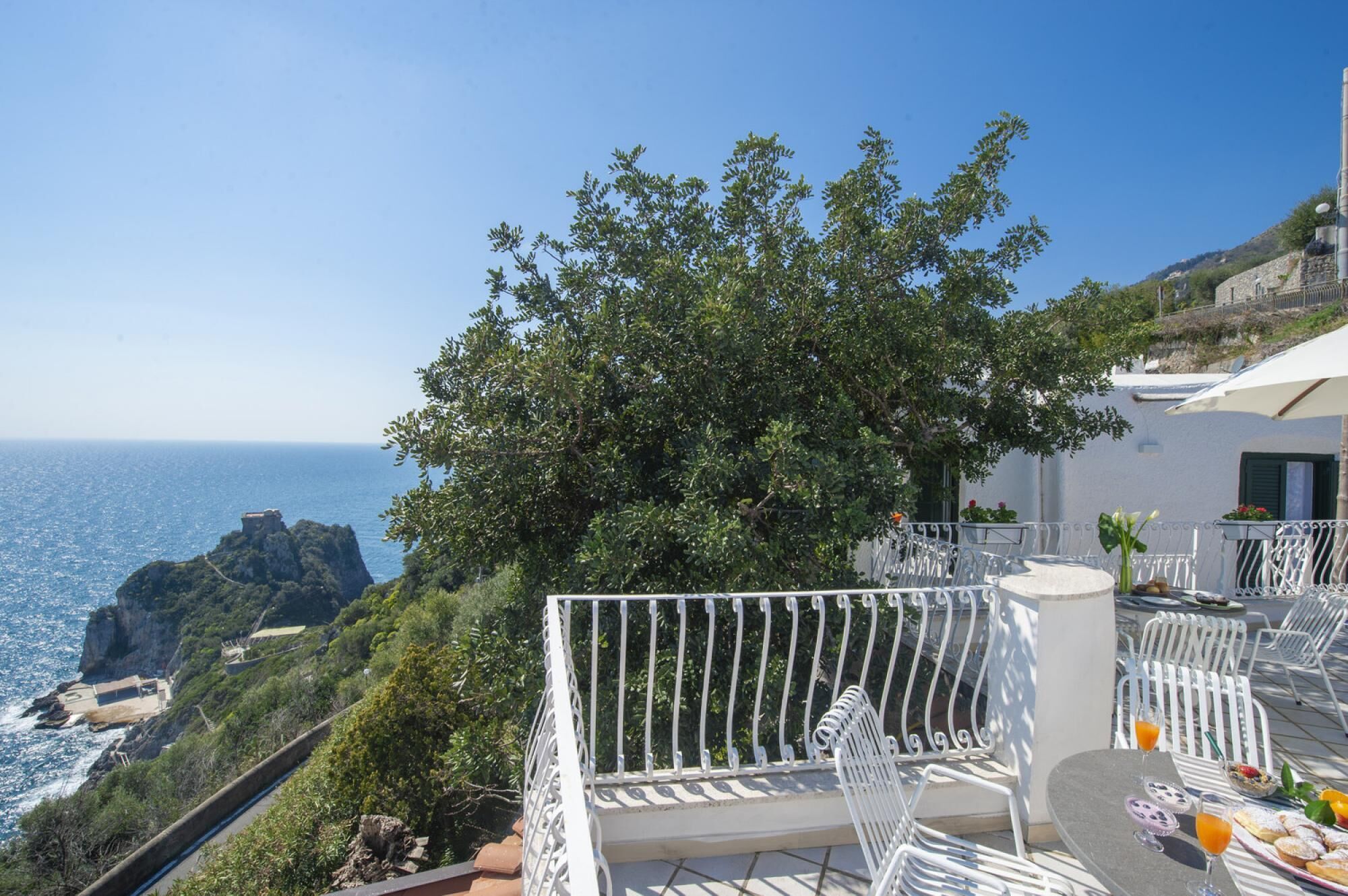 A terrace with white metal chairs and a table, overlooking a coastal landscape with cliffs and the sea.