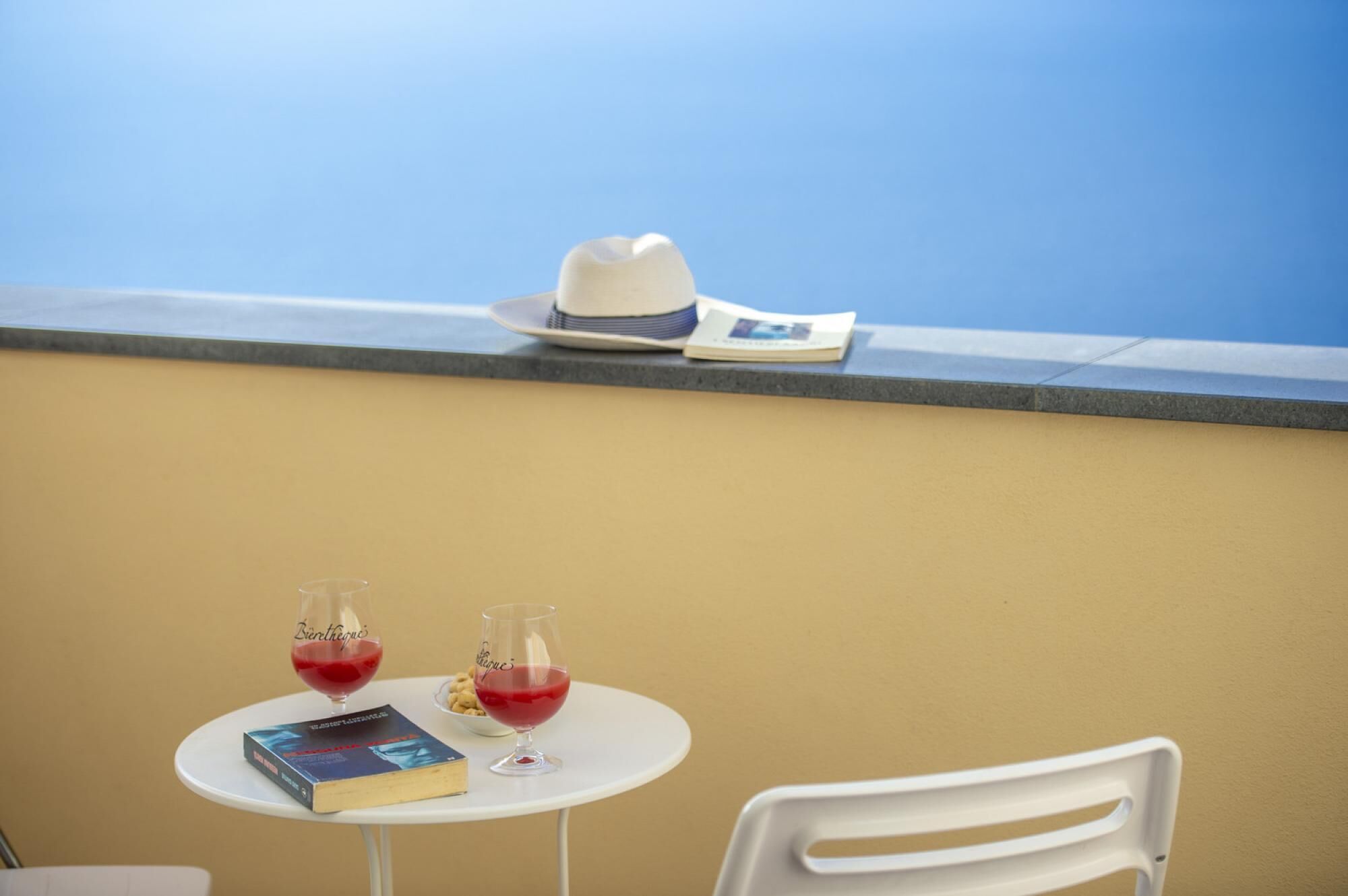 A table with two glasses of red drink, a book, and a white hat on a balcony with a blue sky view.