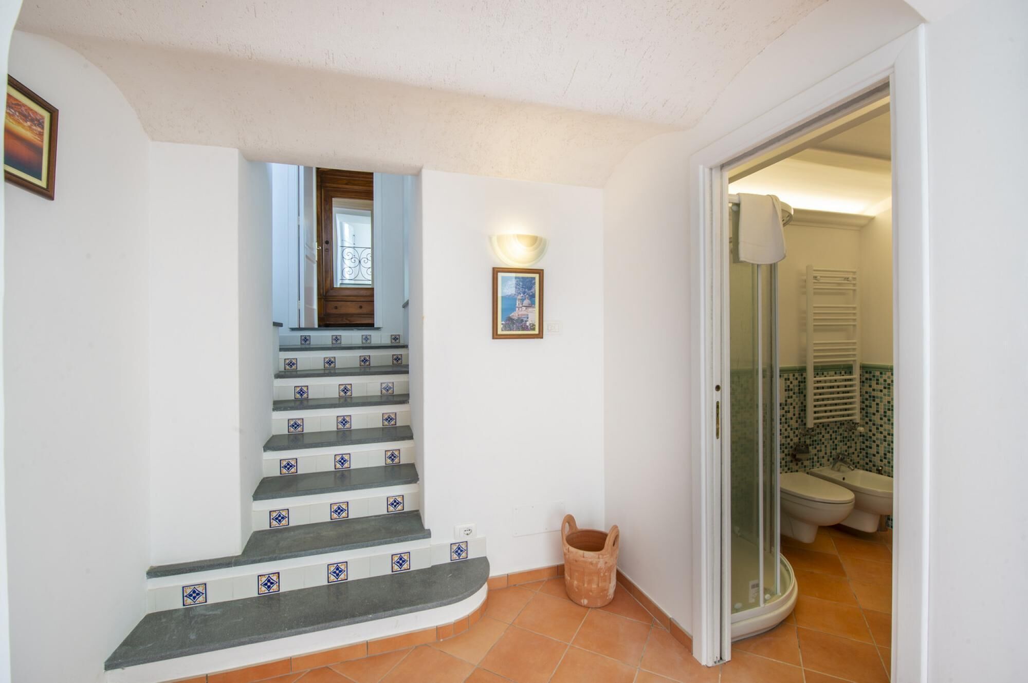 Un escalier orné de carreaux bleus et blancs, une porte en bois, un tableau encadré et une porte de douche en verre donnant sur une salle de bain dont le sol est carrelé.