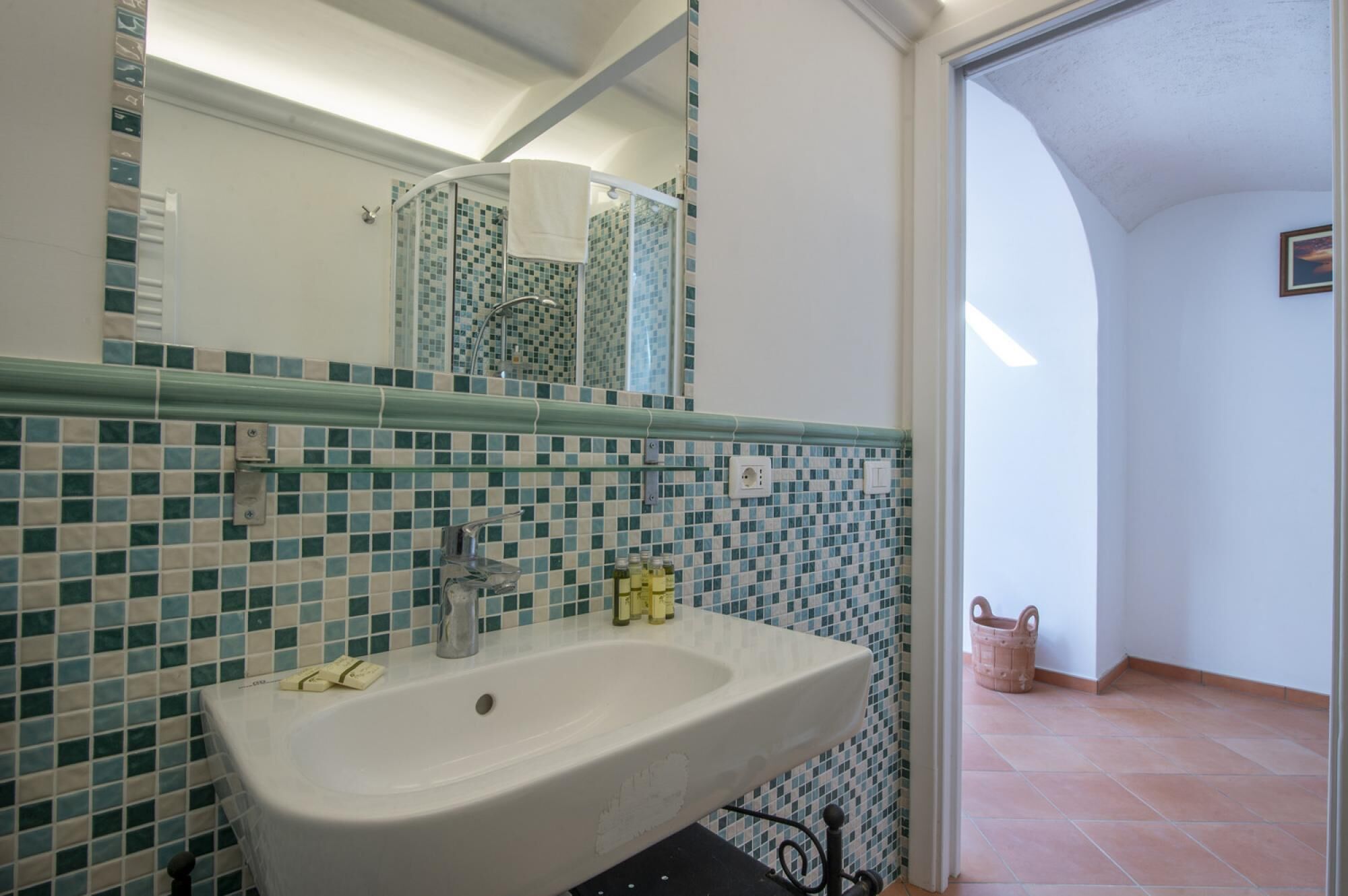 A bathroom with a white sink, green and blue mosaic tiles, a glass shower enclosure, and a visible door leading to another room.
