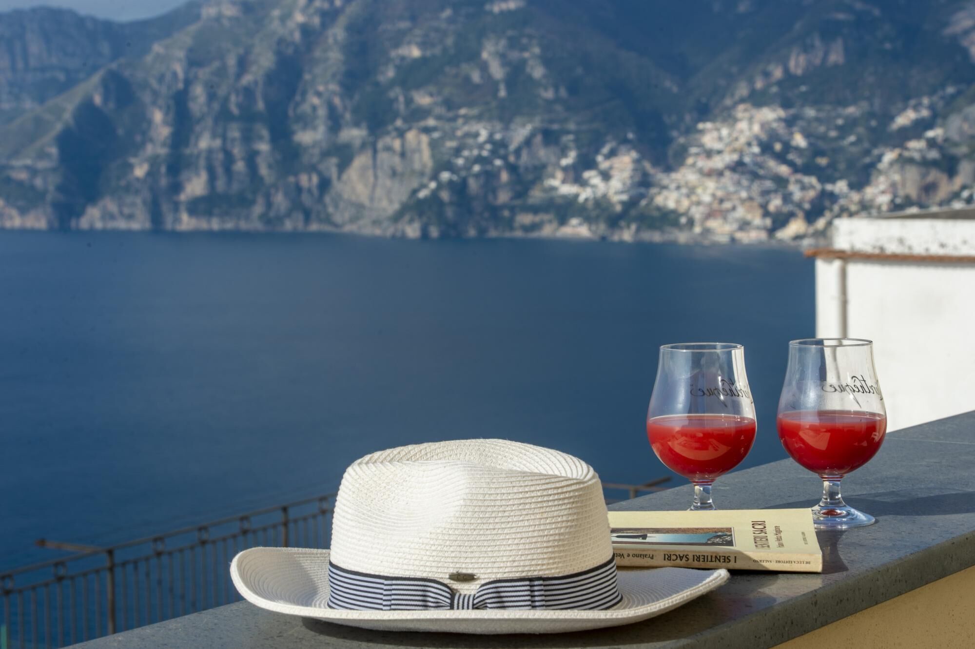A white straw hat, two glasses of red drink, and a menu on a balcony overlooking a body of water and mountains.