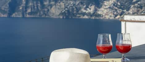 A white straw hat, two glasses of red drink, and a menu on a balcony overlooking a body of water and mountains.