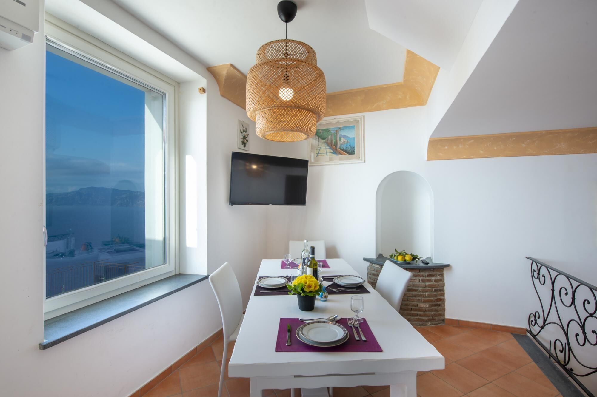 A dining area with a table set for two, a large window with a view, a television mounted on the wall, and a hanging woven light fixture.