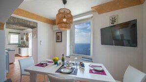 A dining area with a table set for four, a large window with a view of the sea, and a flat-screen TV mounted on the wall. - Villa Cimino - Central Villa Stunning Sea View (Praiano)