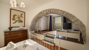 A bedroom with a stone archway, a bed with rolled towels, a dresser, a painting, and a television in the adjacent room. - Villa Cimino - Central Villa Stunning Sea View (Praiano)