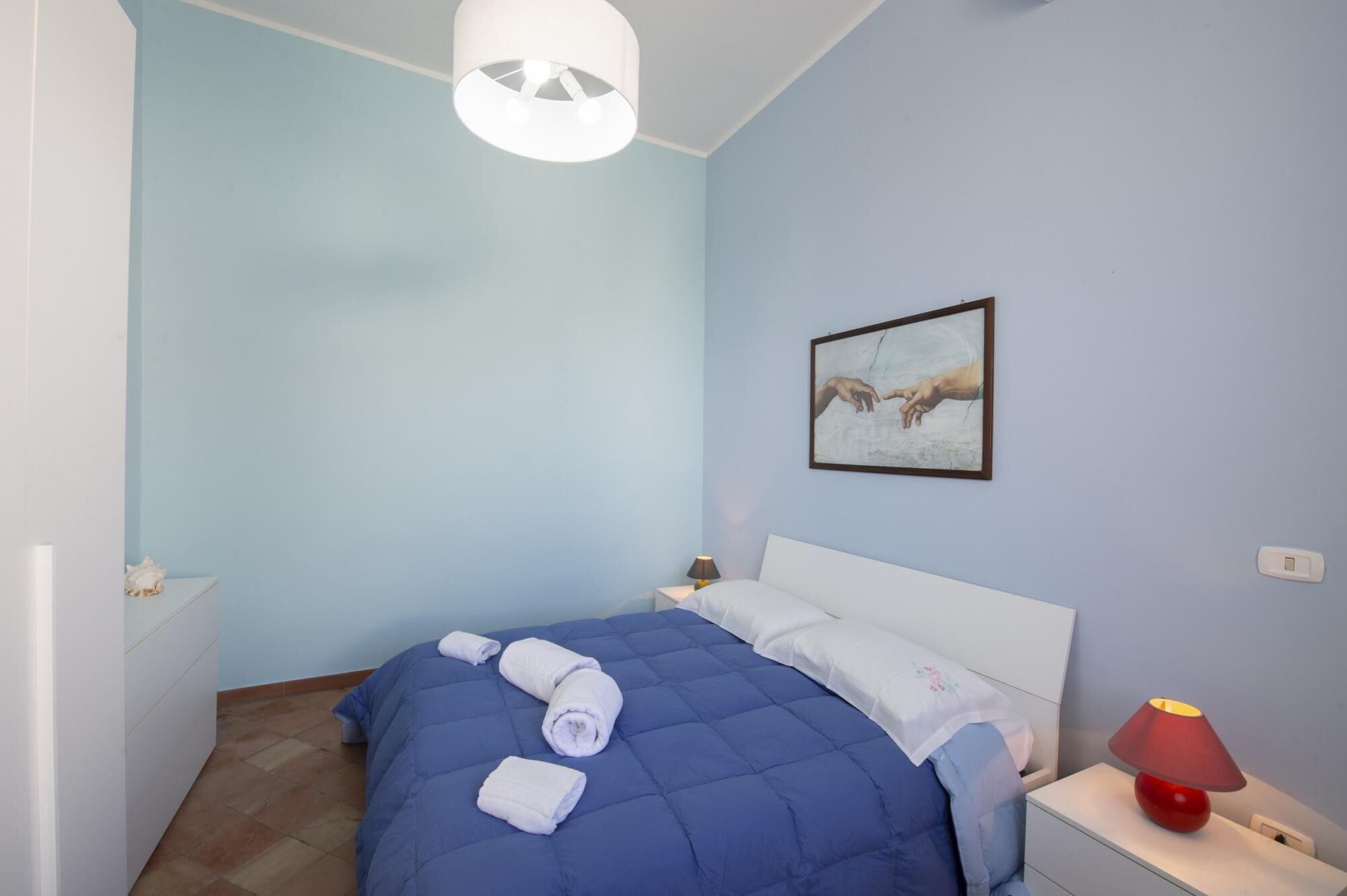 A bedroom with a blue bedspread, white pillows, a framed artwork on the wall, a bedside lamp, and a red lamp on the nightstand.