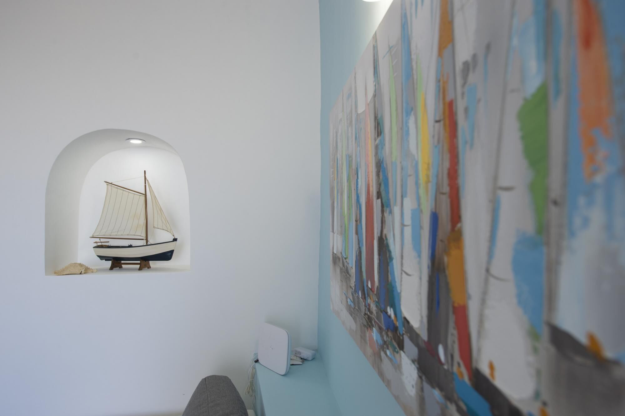 Un velero modelo en un hueco, una pared blanca y un cuadro abstracto y colorido.