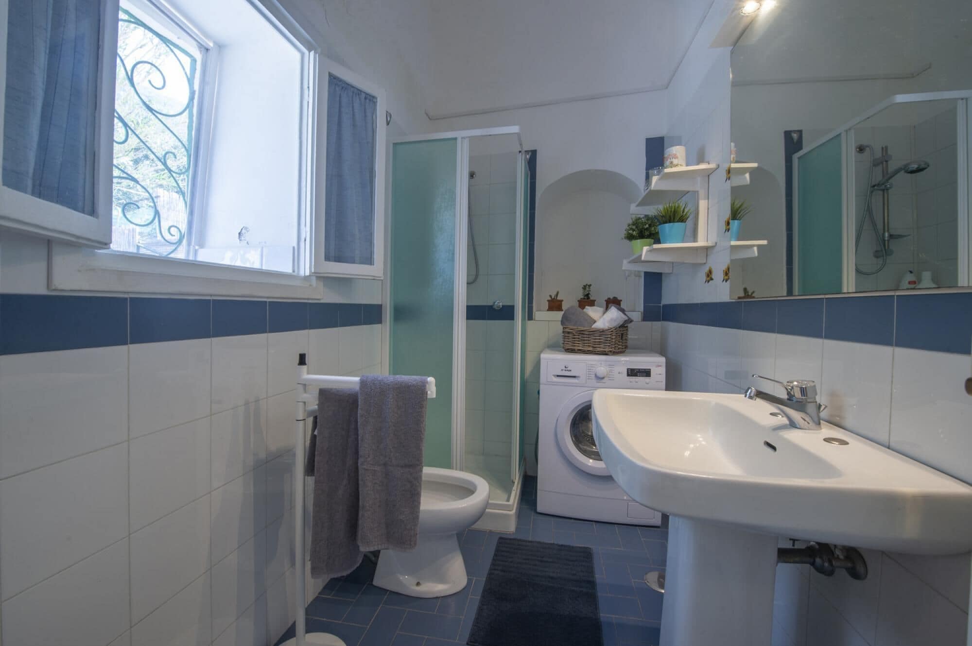 A bathroom with a toilet, washing machine, sink, and a window with curtains.